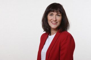 A woman with dark hair, she is looking into the camera and smiling. 