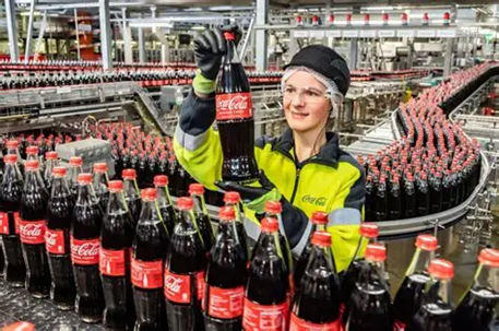 A woman standing next to a conveyor belt full of Coca Coca bottles. She is holding a bottle of Coca Cola. 