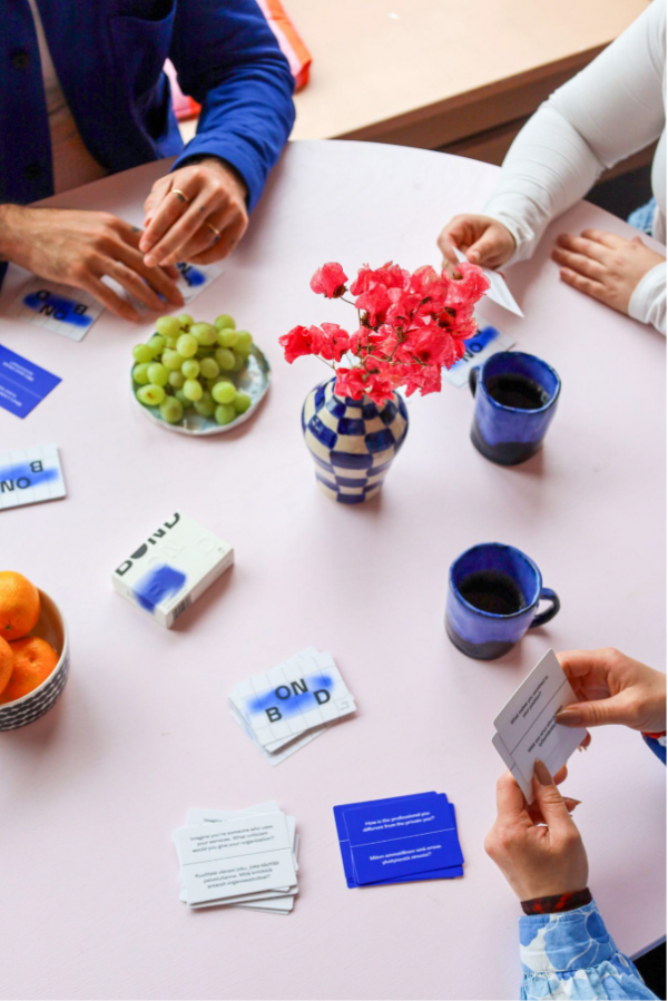 A table with coffee cups and fruit, you see the hands of the people sitting around the table, playing a card game.