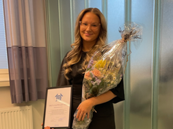 En kvinna håller i en blombukett och ett diplom, ser in i kameran och ler. 