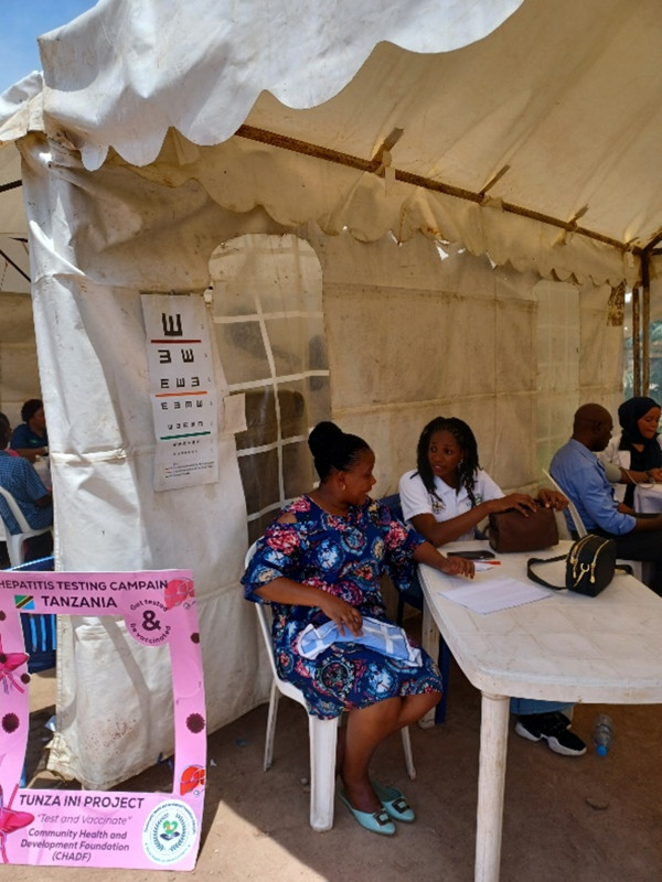 Personer som sitter runt bord i ett vitt tält. I förgrunden syns en skylt med texten Hepatitis Testing Campain Tanzania. Get tested & be vaccinated. Tunza Ini Project.