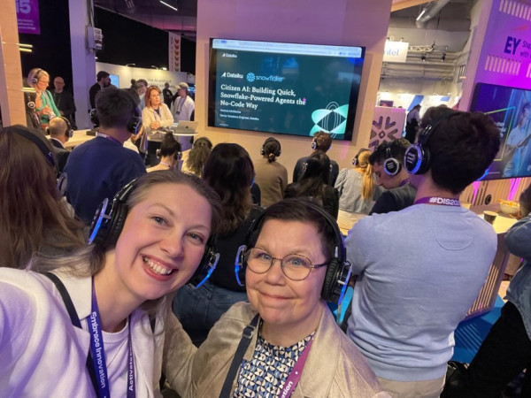 En selfiebild med två kvinnor som ser in i kameran och ler. De har båda på sig hörlurar. I bakgrunden syns en folksamling och en TV-skärm med en presentation med namnet Citizen AI: Building quick, snowflake-powered agents the no-code way.