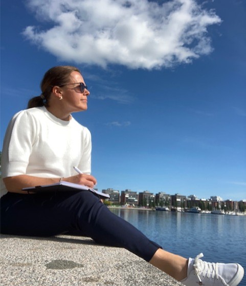 En kvinna sitter vid ett vattendrag och tittar bort från kameran. Hon har ett skrivblock i famnen, en penna i handen och solglasögon.
