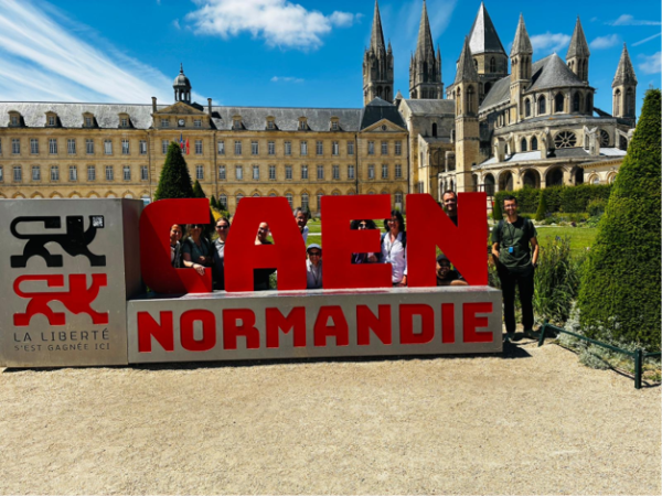 En bild av ett slott. På en grusplan framför slottet finns en stor skylt med texten CAEN - NORMANDIE. Bredvis skylten står en man.
