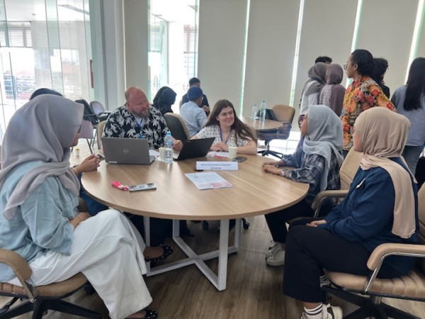 A picture of people, 6 women and one man, workshopping around a round table. Two people are working on laptops. All people around the table are talking to each other.