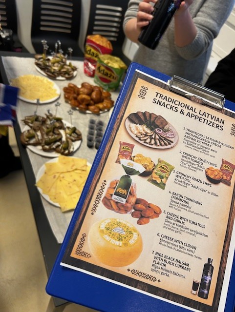 I bakgrunden av bilden syns ett bord med uppdukad mat. I förgrunden syns en skylt med texten Tradicional Latvian Snacks & Appetizers